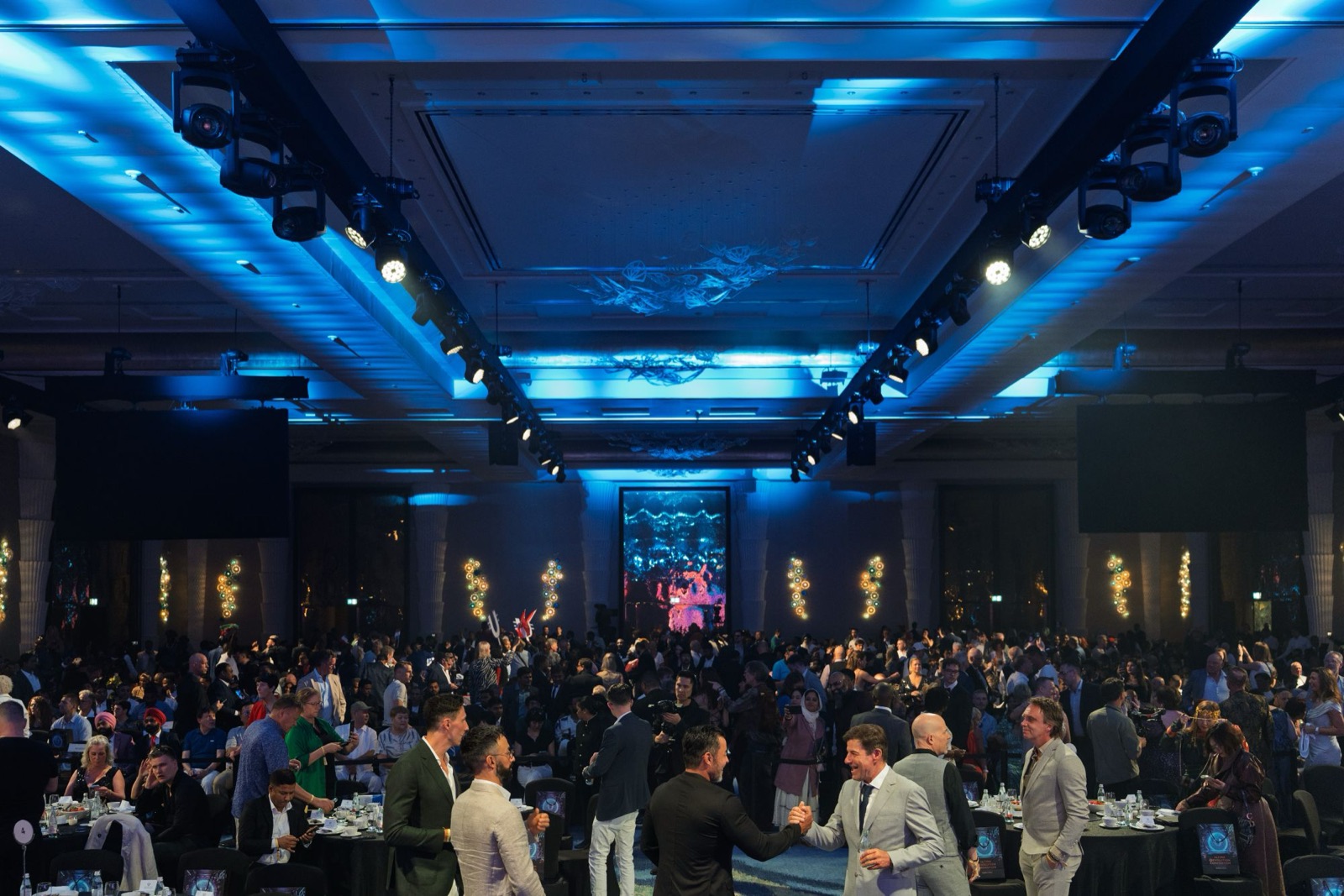 Corporate event photo coverage: a gala dinner in a ballroom under blue lighting, guests greeting each other between the tables