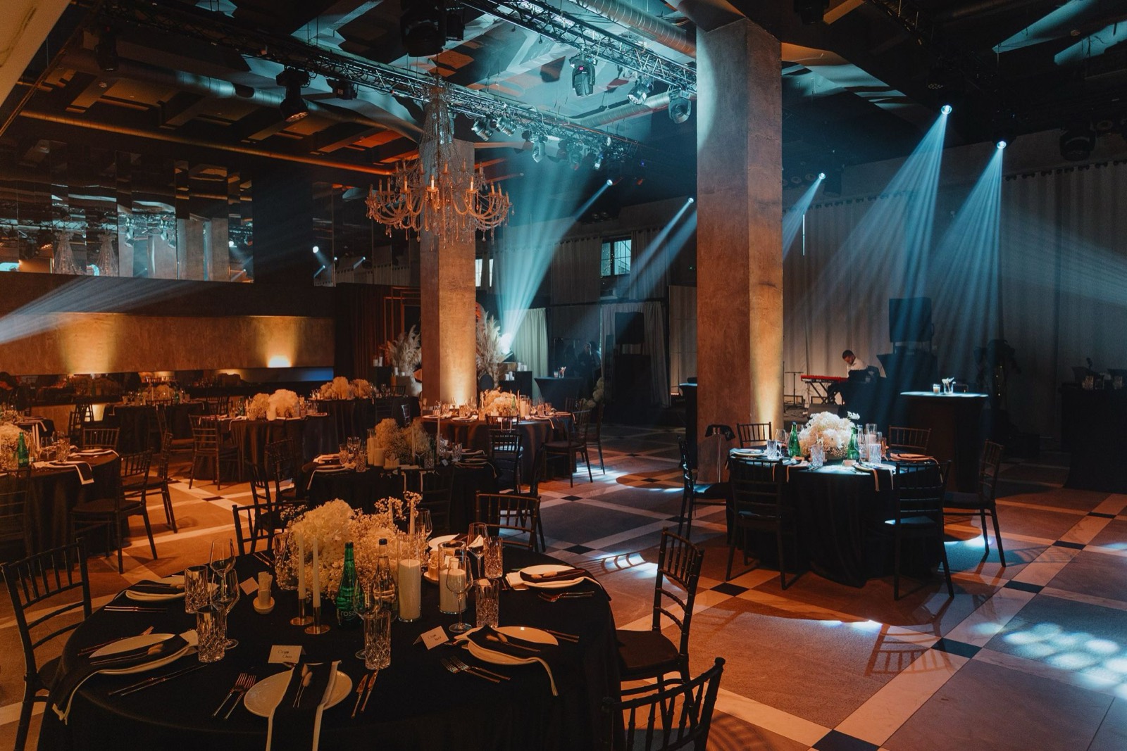 Private corporate gala for 60 guests in Belgrade: a ballroom set for dinner, round tables in black with white flowers and candles, a crystal chandelier and blue stage lights
