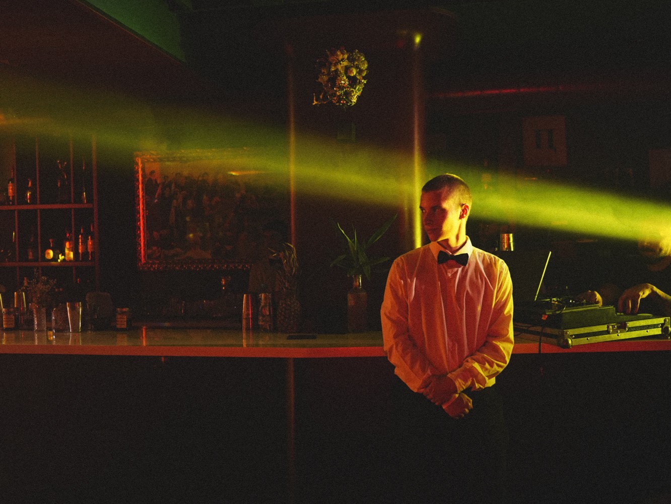 Private party in 1990s style: a waiter in a bow tie by the bar, a green light beam cutting through the dark room