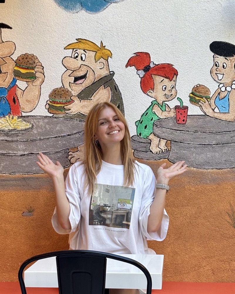 Masha laughing with raised hands in front of a hand-painted Flintstones mural