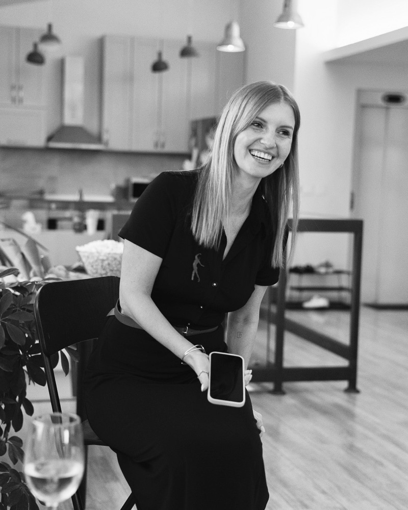 Black-and-white photo of Masha laughing on a chair in a bright loft, phone in hand