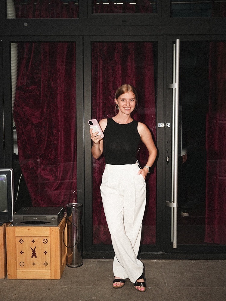Masha Welt, producer based in Belgrade, in a black top and white trousers, smiling, by a glass door with velvet curtains